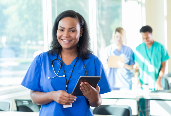 Smiling healthcare worker in blue scrubs with a stethoscope holds a tablet in a bright hospital office, with two colleagues blurred in the background near windows.