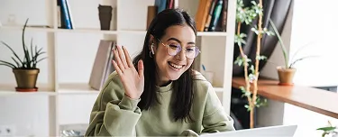 1. A woman sitting at a desk with a laptop, smiling and waving at the camera.
