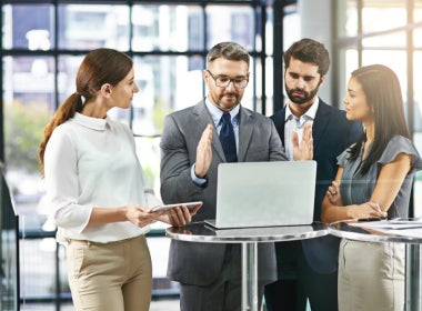 A group of four professionals gathered around a laptop on a table, engaged in a discussion in a modern office setting.