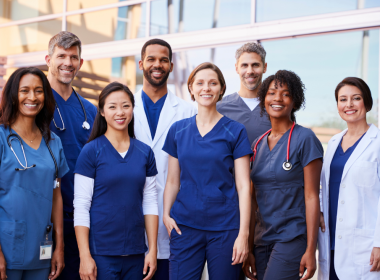 A diverse group of medical professionals smile at the camera while standing outside a building.