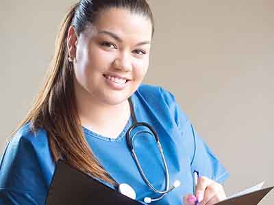 A nurse in blue scrubs holding a clipboard and a pen, with a stethoscope.